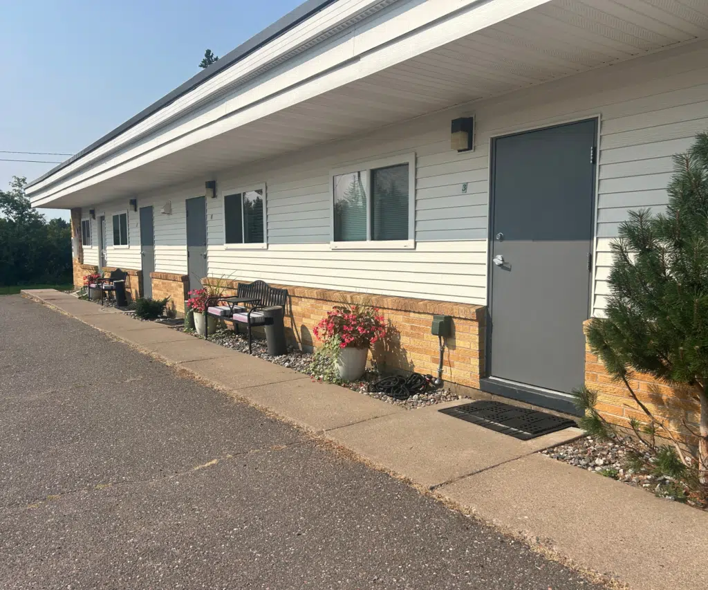 Exterior view of a single-story motel building featuring grey entry doors, outdoor seating, potted flowers along a concrete walkway, and a paved parking lot.