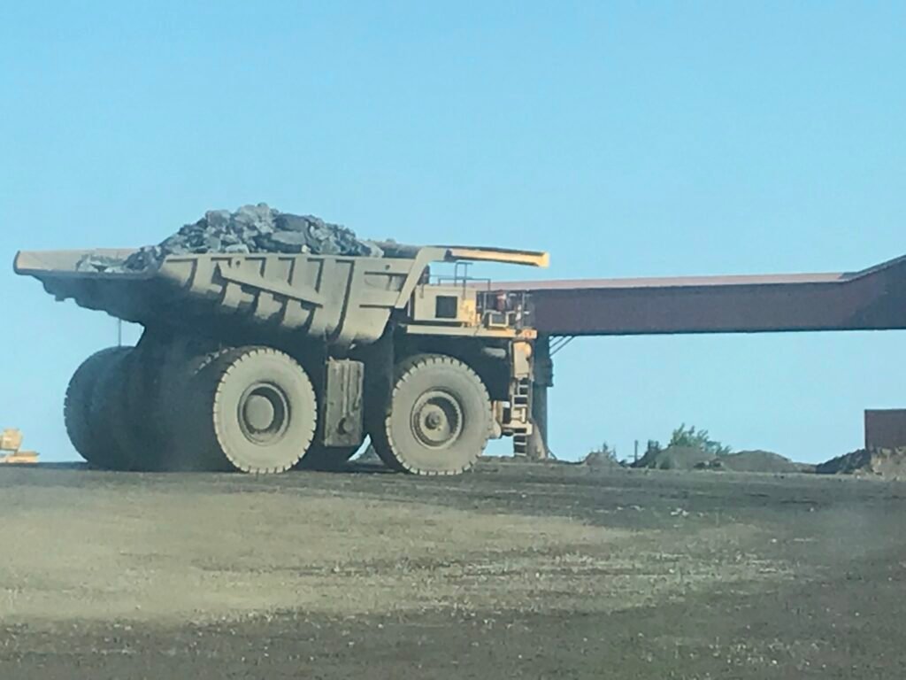 Places to Explore A massive yellow haul truck fully loaded with iron ore driving through an open-pit mine facility under a clear blue sky.
