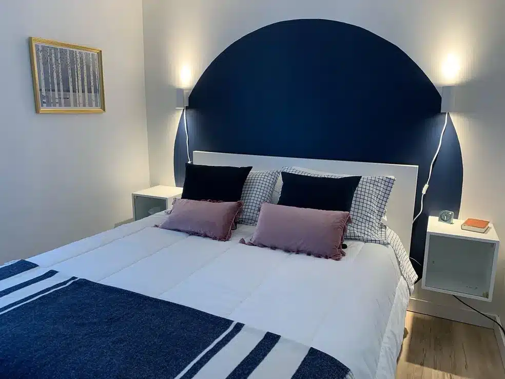 Interior view of Suite 1 bedroom featuring a bed with a blue stripe accent throw, a painted dark blue arch wall feature, floating white nightstands with mounted reading lamps, and framed art on light walls.
